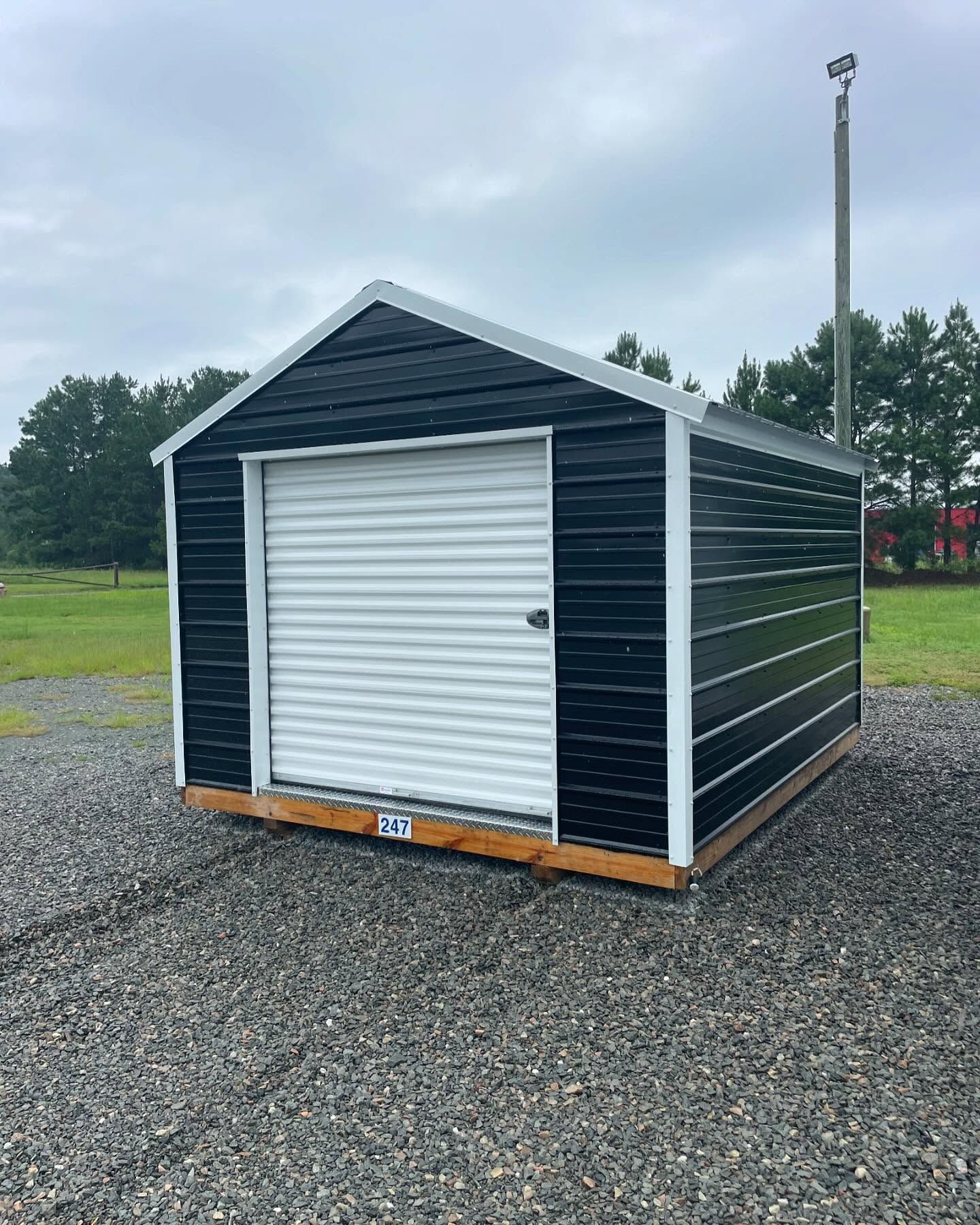 Black storage shed with a white roll-up door on gravel, with a tall pole to the right.