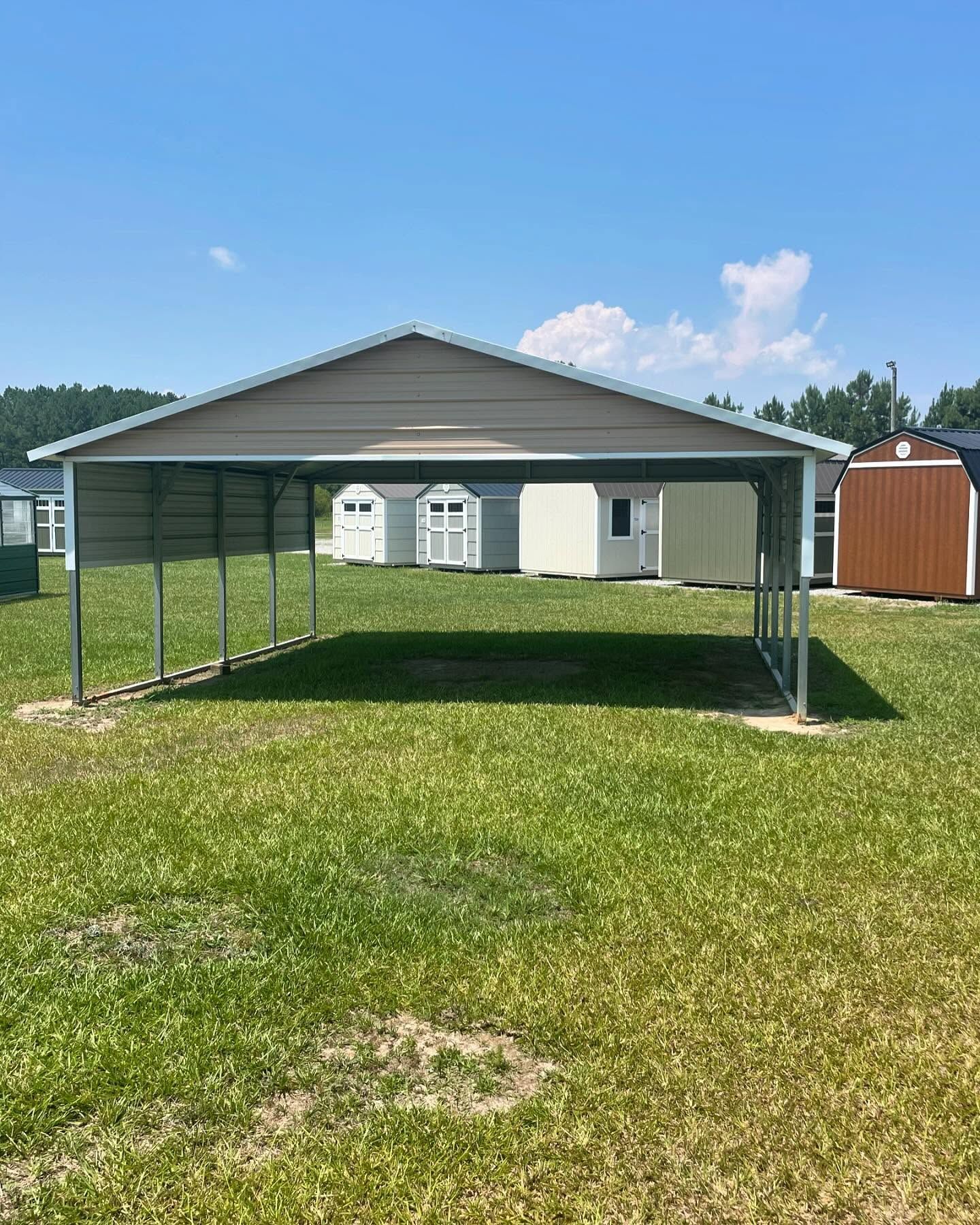 Metal carport in a grassy field with several sheds in the background under a blue sky.