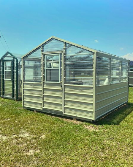 Glass greenhouse in a sunny yard, filled with potted plants on shelves.