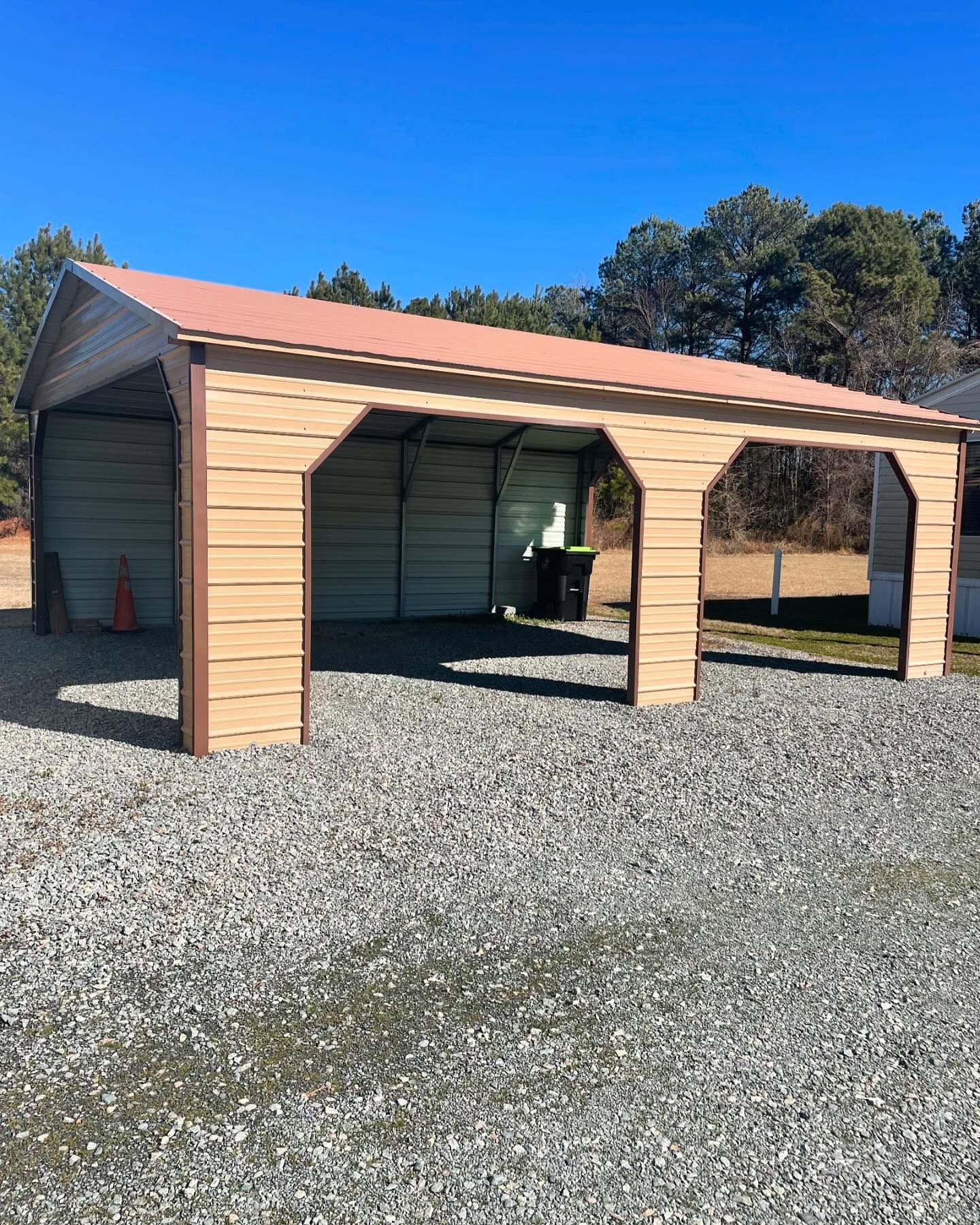 A metal carport with tan beams and brown roof, on gravel ground, under a clear blue sky.