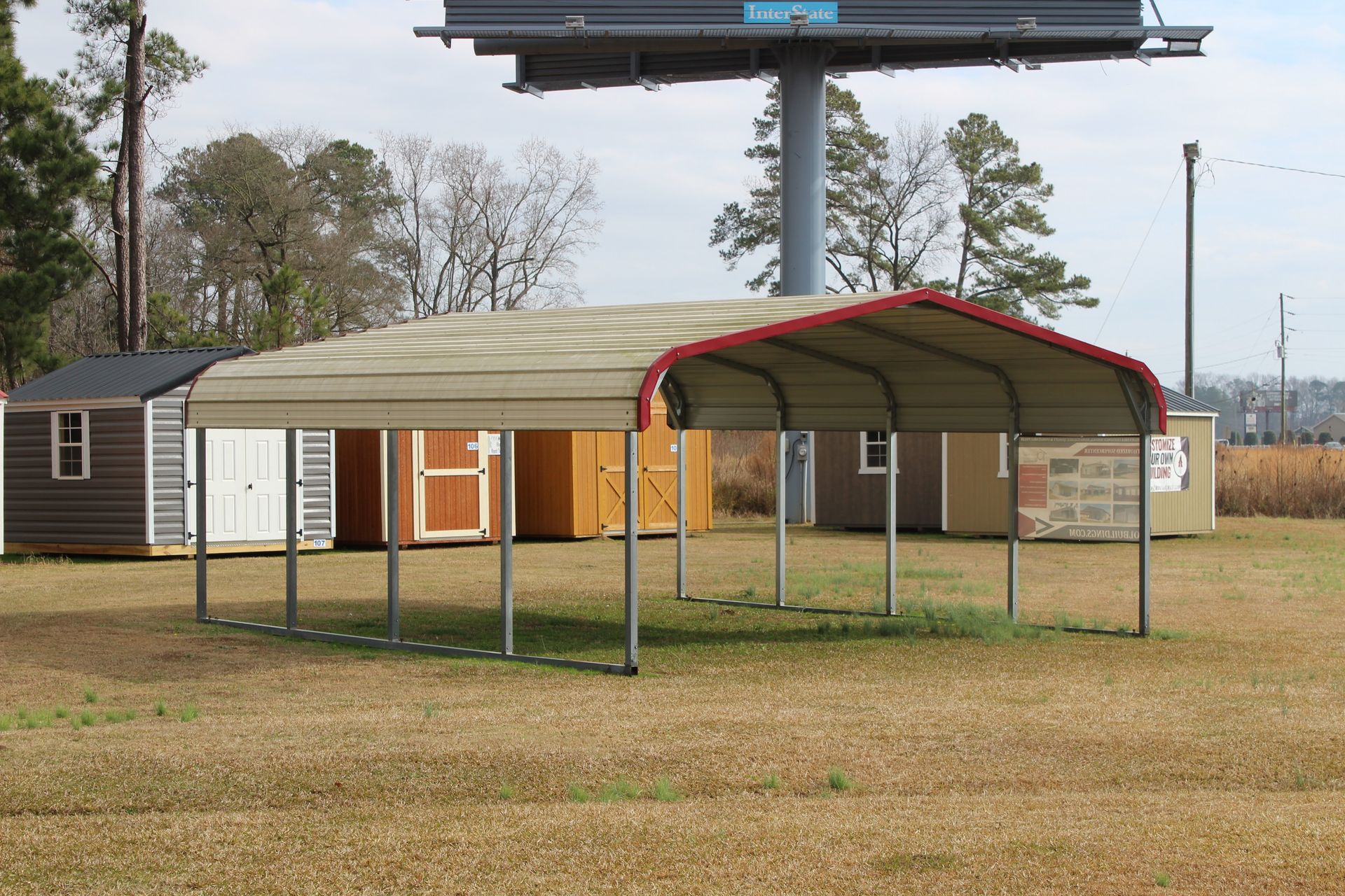 Carport with red trim in a grassy field, with multiple sheds in the background.