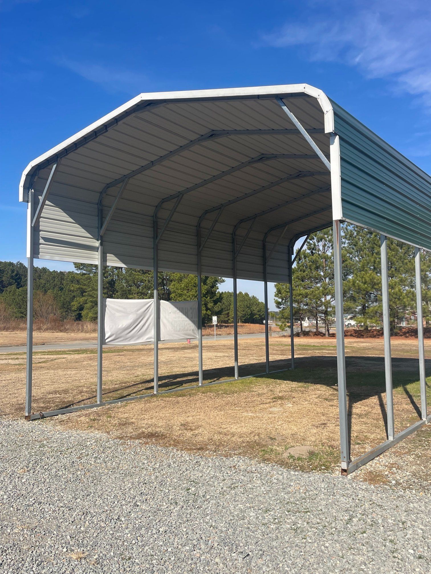 Metal carport with a green roof, on a gravel surface under a bright blue sky.