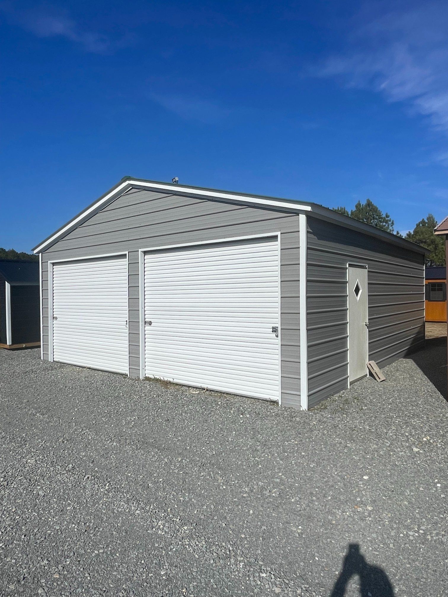 Gray two-car garage with white doors and a side door, set on gravel. Clear blue sky in the background.