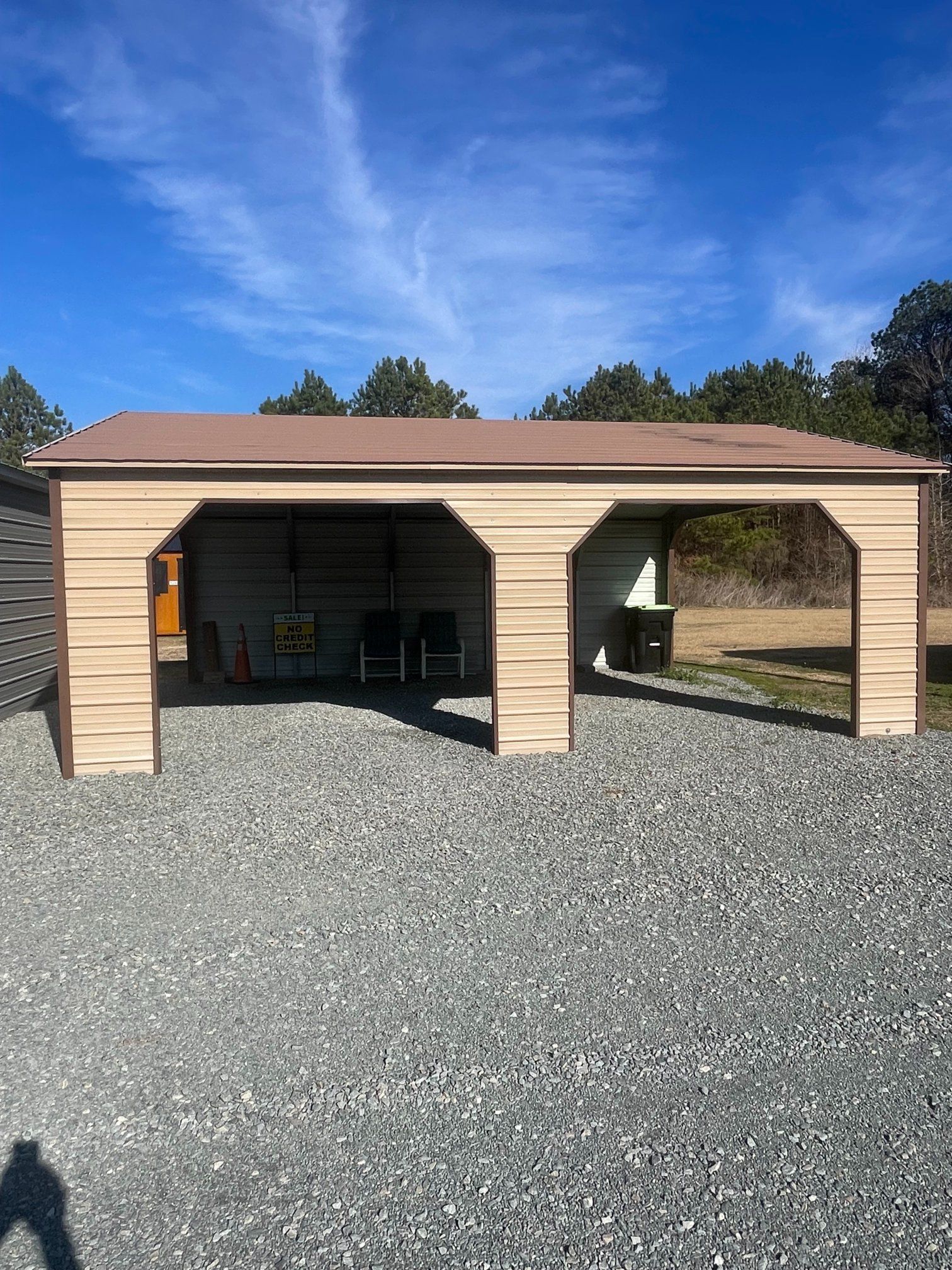 Tan metal carport on gravel with brown roof, under a blue sky.