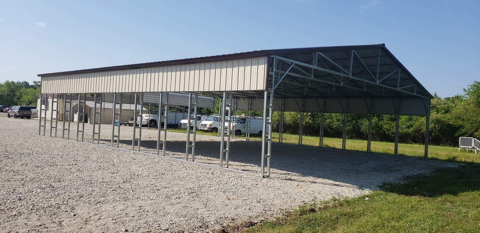 Metal carport with cars parked underneath. Gravel ground and green grass. Blue sky in background.