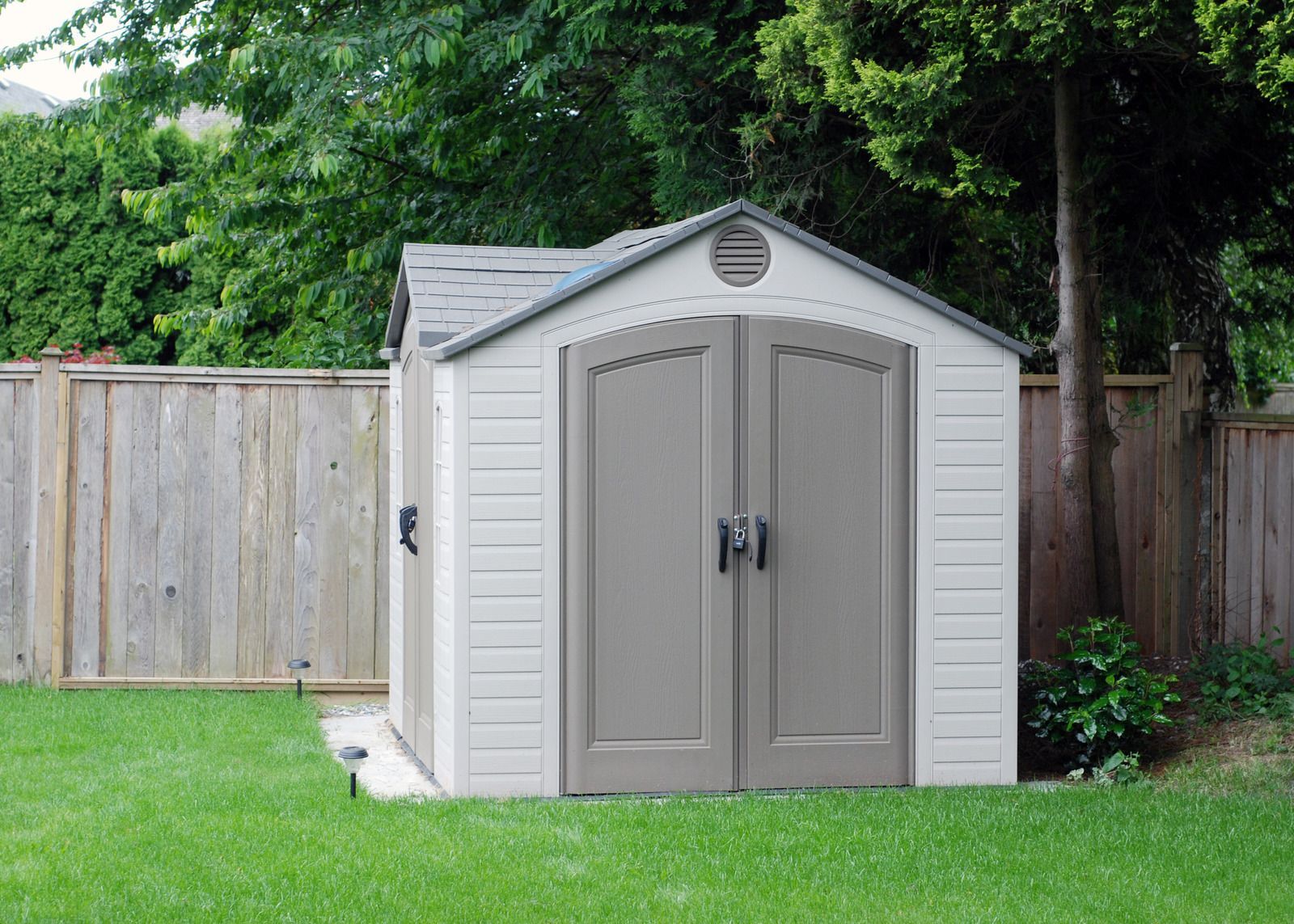Tan shed with gray doors and roof, in a backyard setting near a wooden fence and green trees.
