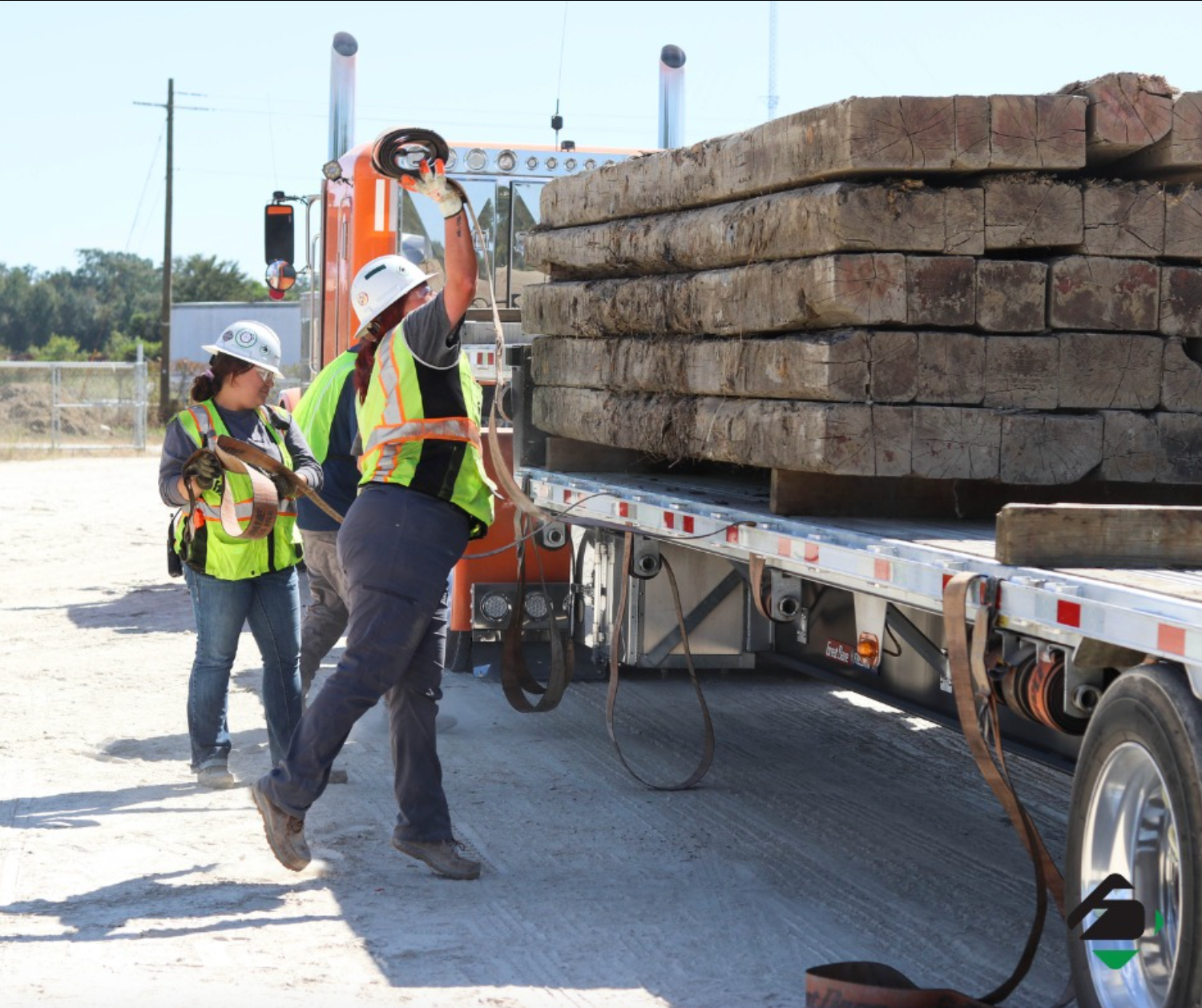 Two women working to throw ropes across flatbed truck with lumber loaded on. 