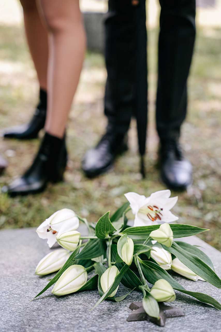White Lily Bouquet Resting On A Grave Marker With Mourners Standing Nearby In A Cemetery Setting