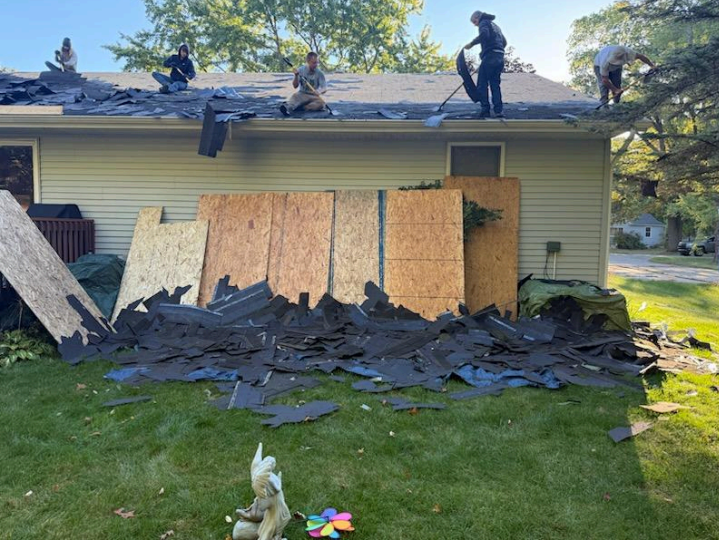 Roofers remove old shingles from a house roof on a sunny day. Debris piles on the lawn.