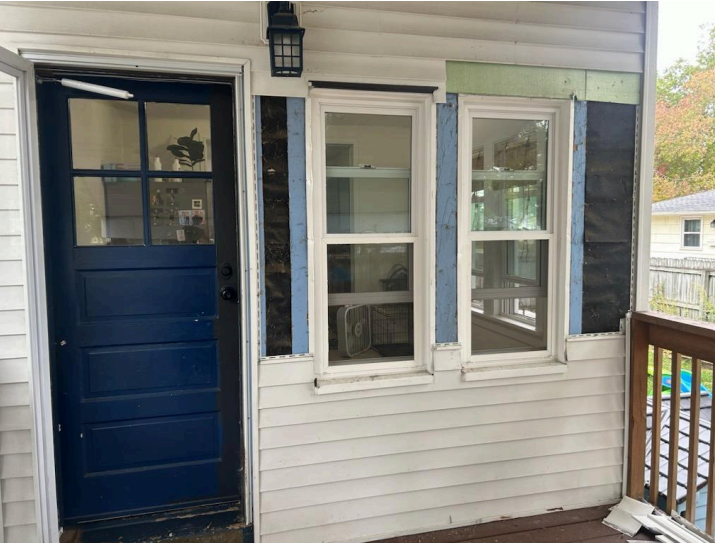 Blue door open next to two white windows, part of siding removed. A porch is to the right.