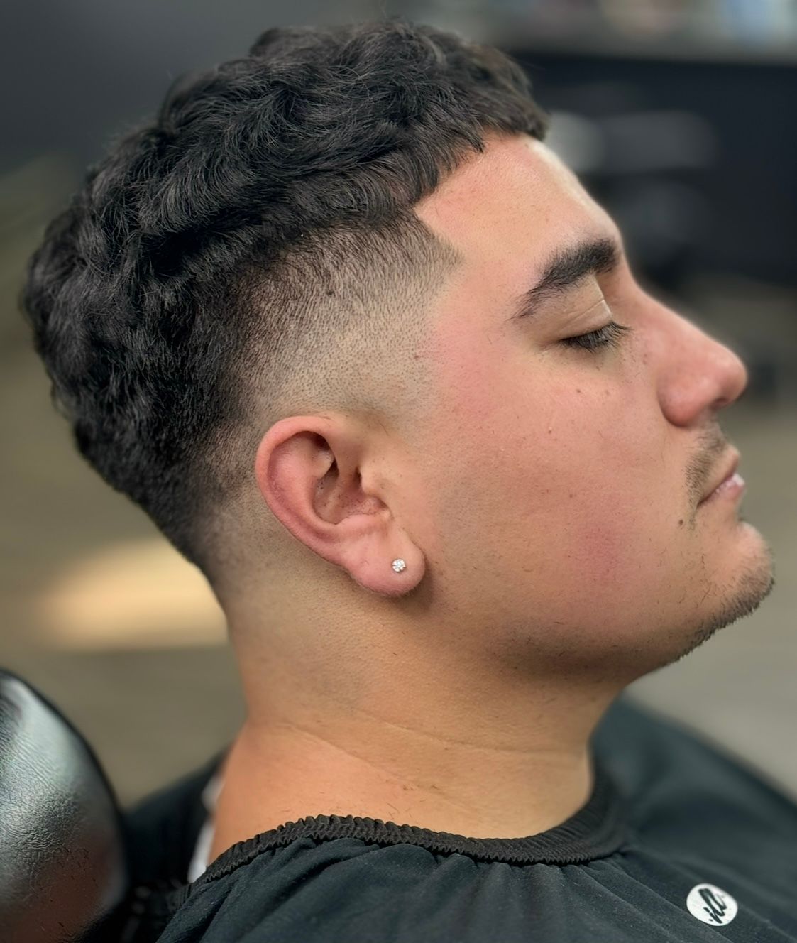 Man with a short haircut, faded sides, and textured curls. He has a slight smile, and is wearing a black cape.