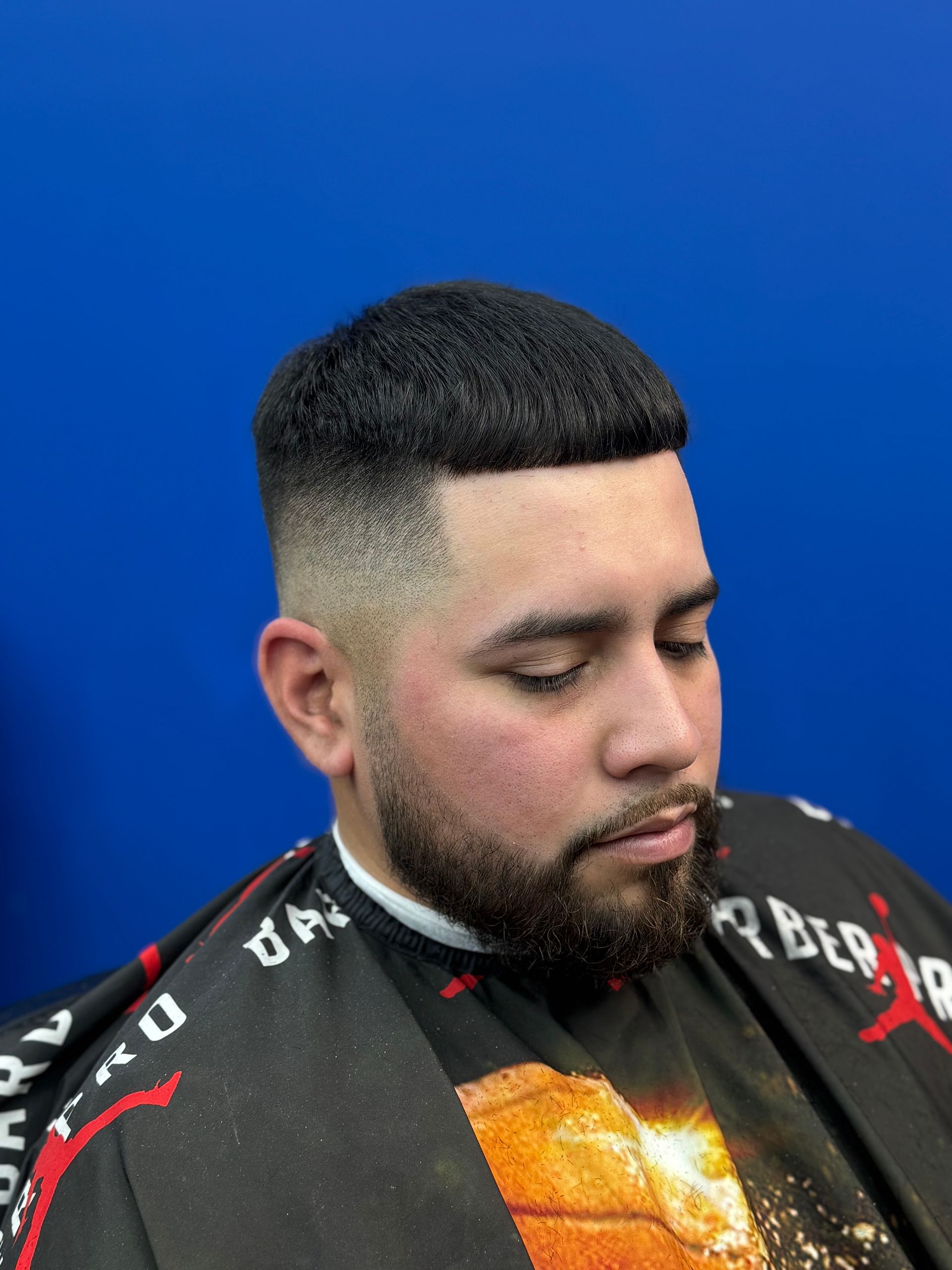 Man with a tapered haircut and beard, sitting against a blue backdrop, wearing a barber's cape.