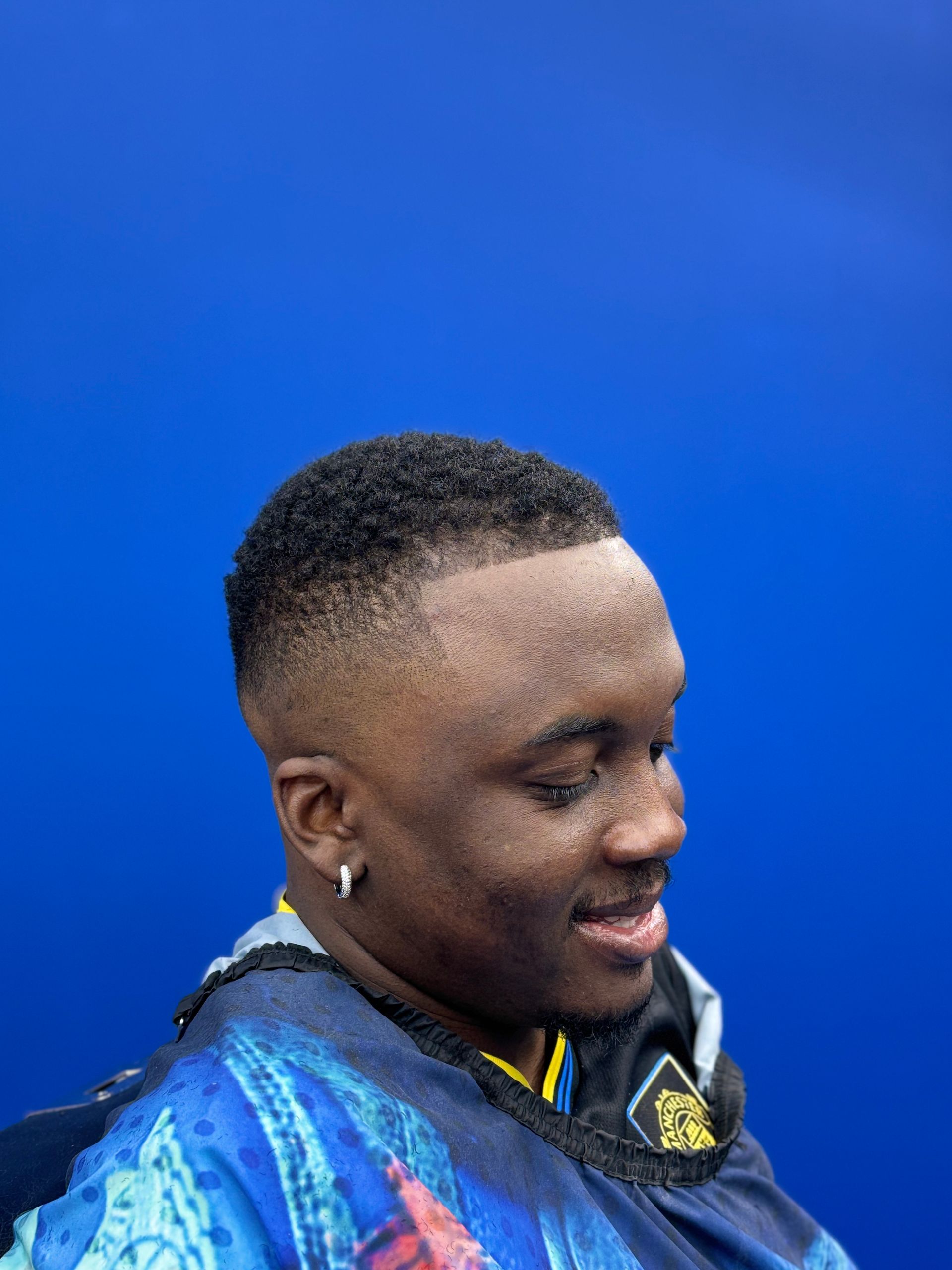 Man with a faded haircut smiling, wearing a blue and yellow patterned jacket, set against a blue backdrop.