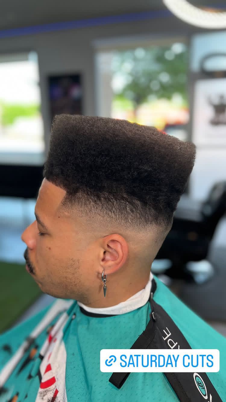 Man with high-top fade haircut, side view. Faded sides, dark top. Wearing a barber cape.