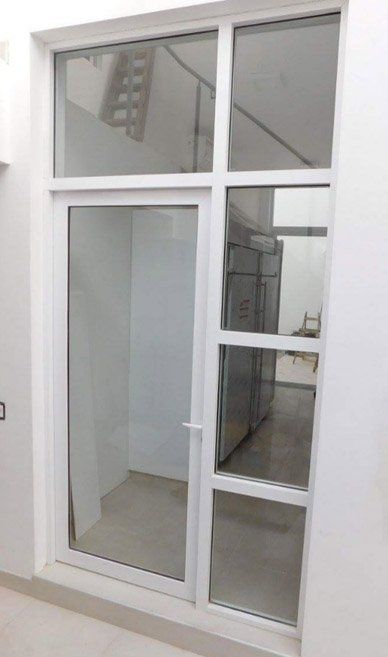 Puerta de cristal con marco blanco y ventanas laterales. La vista interior muestra un largo pasillo y escalera, con paredes de tonos neutros.