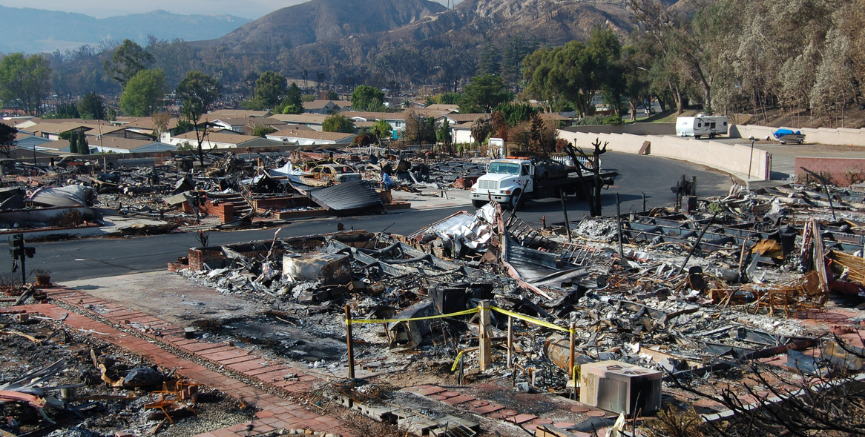 Burned-out homes and debris from a fire, with a truck and mountain in the background.