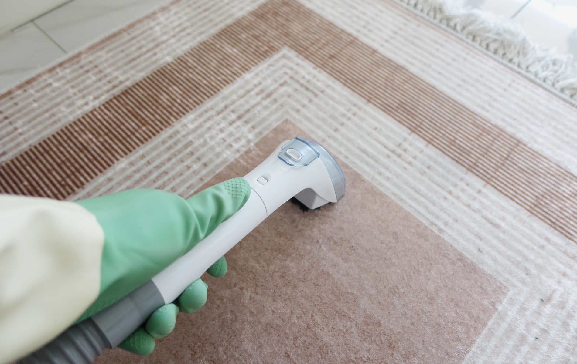 A gloved hand uses a carpet cleaner on a brown and cream patterned rug.