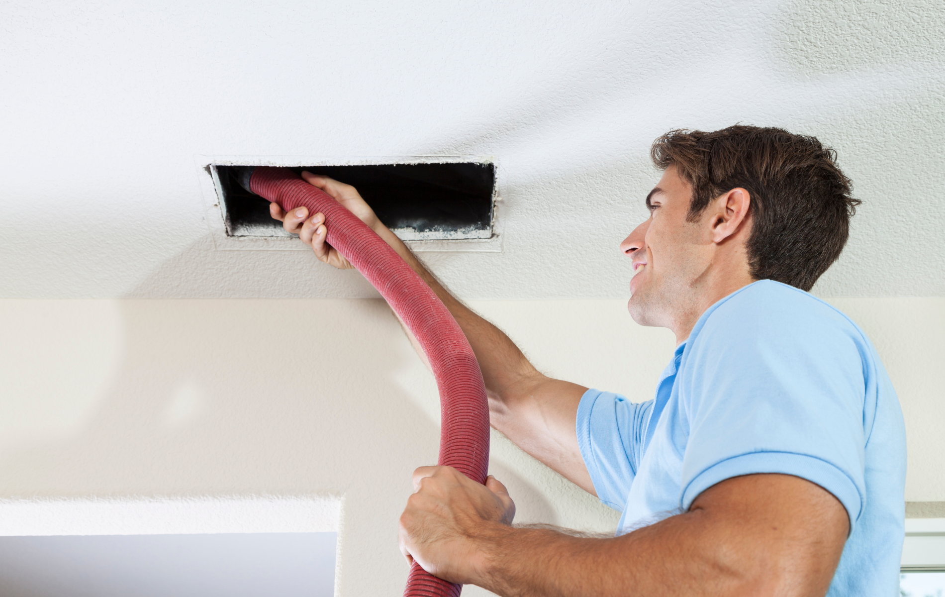 Man cleaning a ceiling air vent with a red hose.
