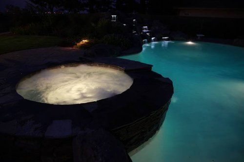 A hot tub is sitting next to a swimming pool at night.