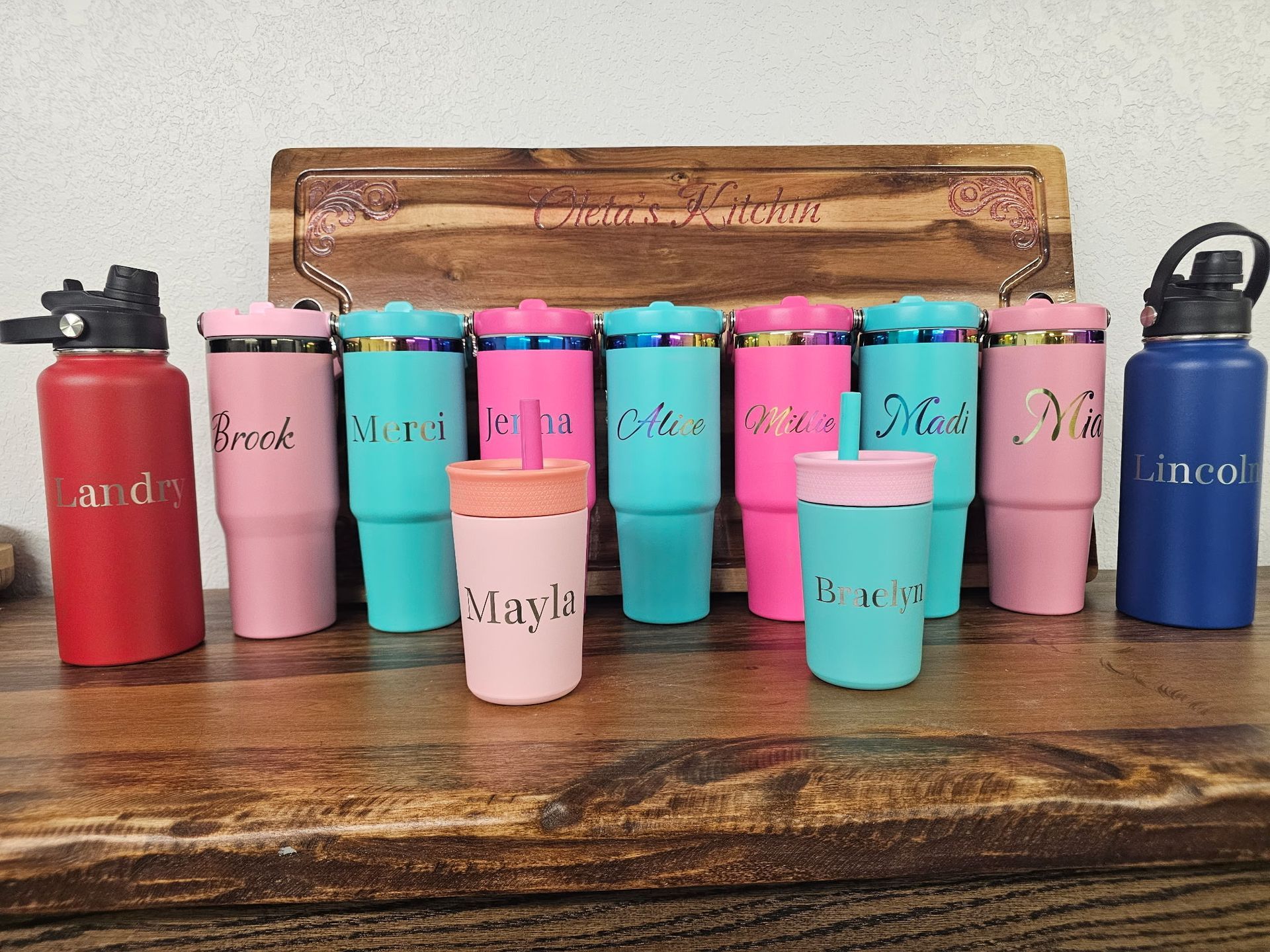 A row of colorful personalized tumblers and water bottles sits on a wooden board, with two smaller cups in the front.