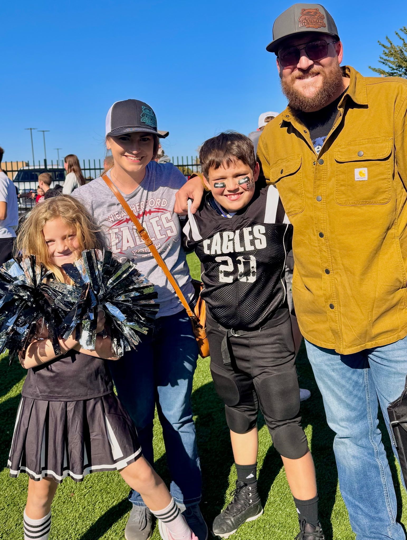 A family posing together at an outdoor sports field; one child in a cheer uniform, another in a football jersey.