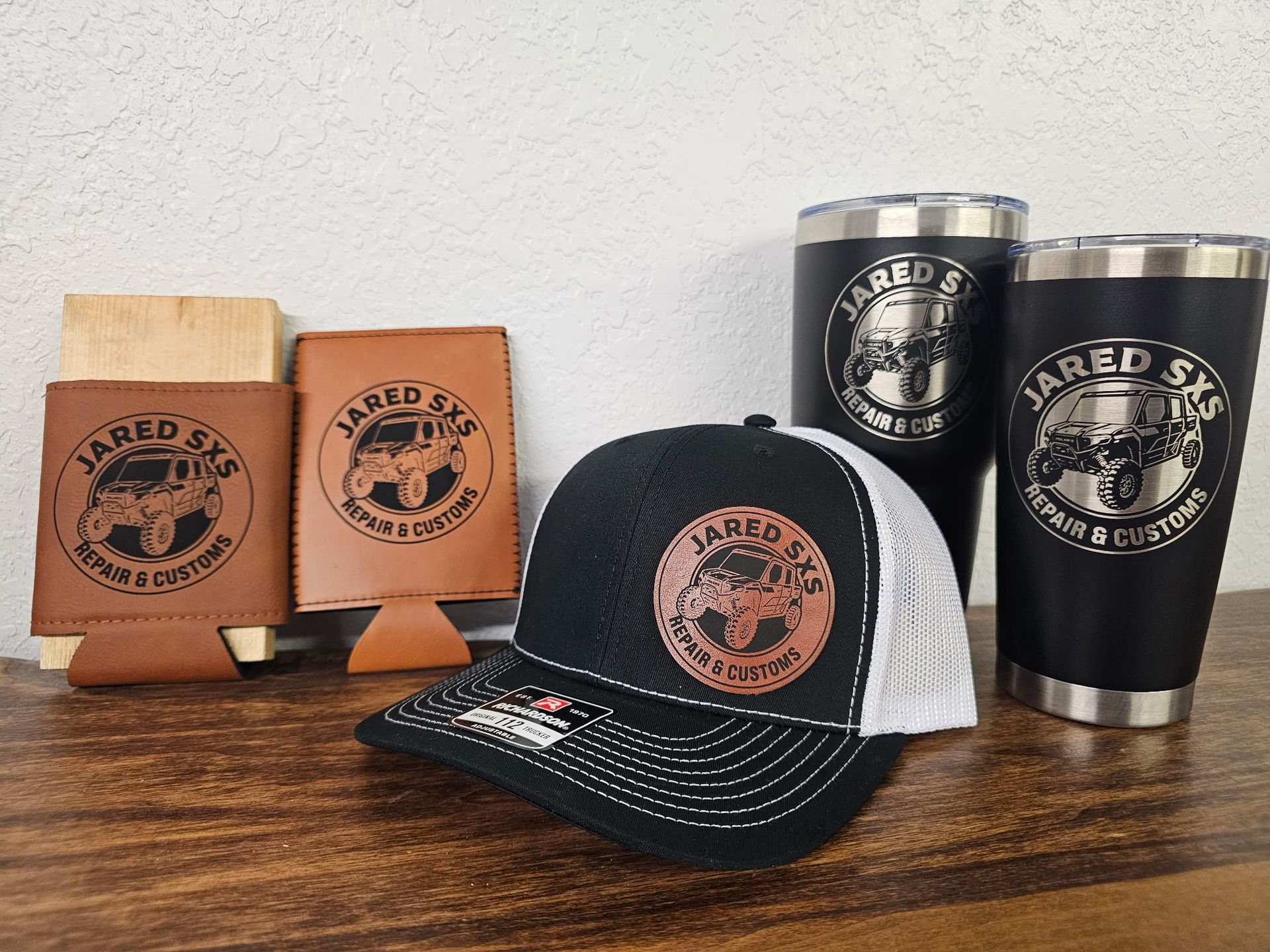 Branded merchandise including two leather koozies, a black and white trucker hat, and two black tumblers on a wooden table.