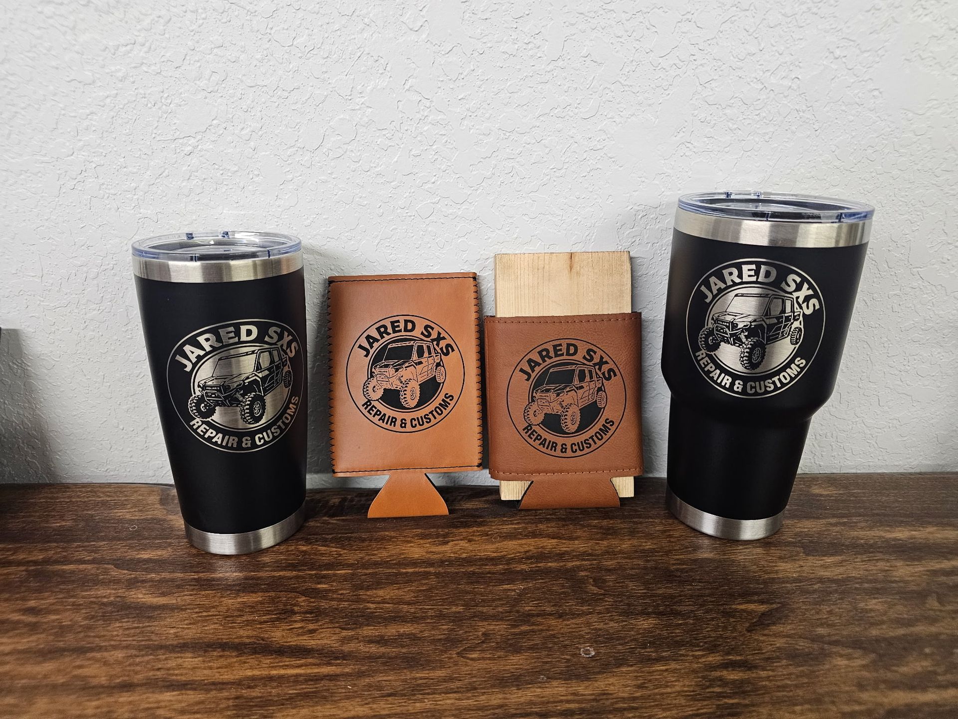 Two black tumblers and two tan leather koozies with a logo on a wooden surface.