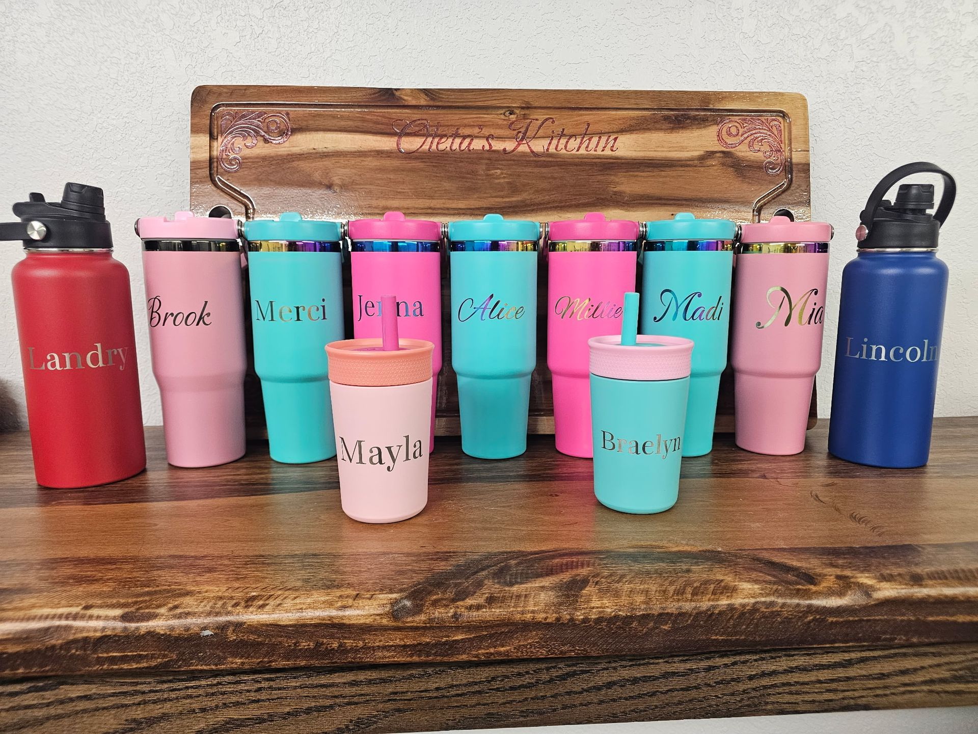 A row of colorful, personalized tumblers and two water bottles with names engraved, displayed on a wooden surface.