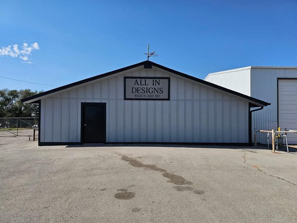 The white, single-story All In Designs shop building with a black door and sign sits under a clear blue sky.