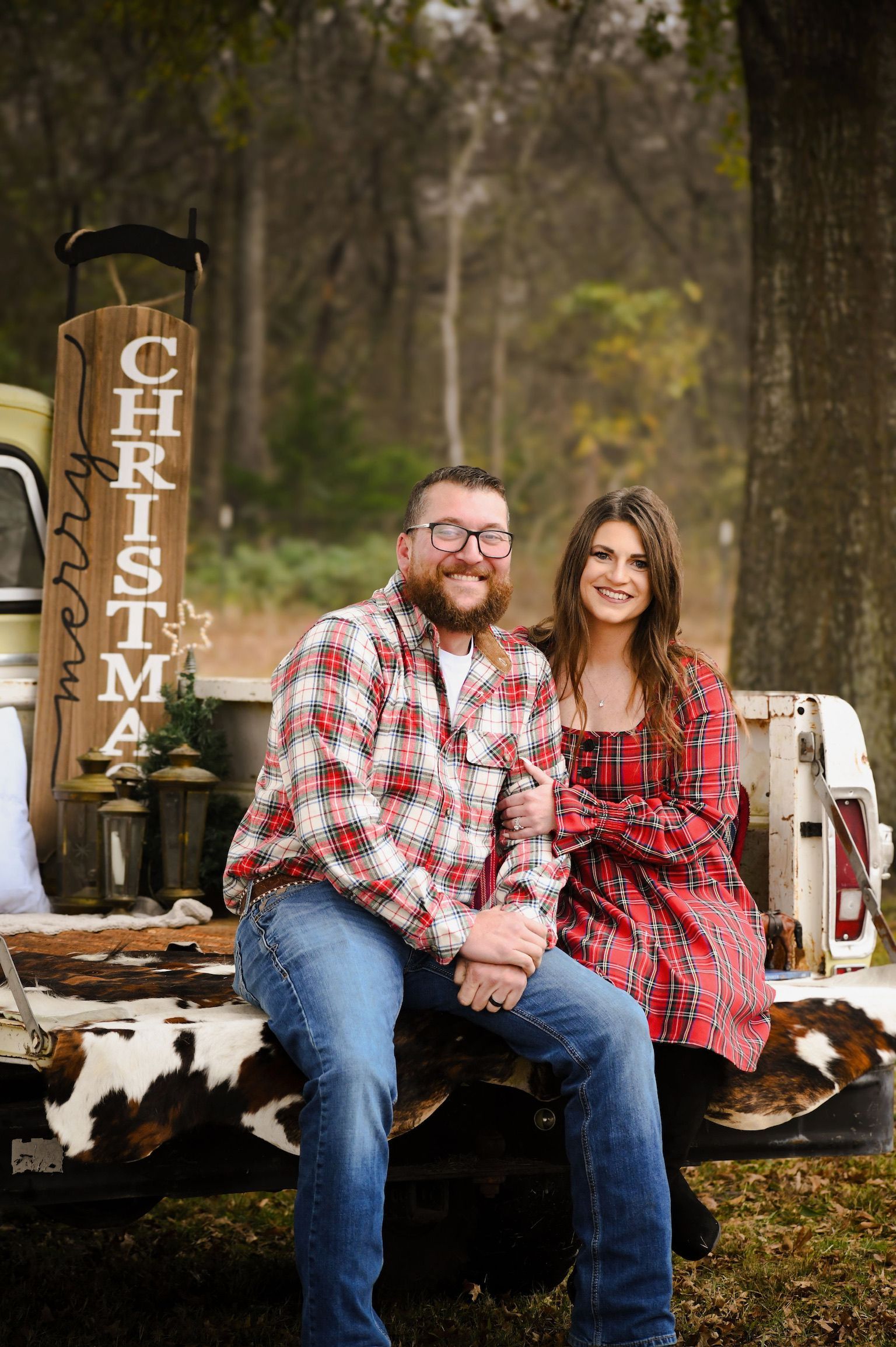 A smiling couple in festive red plaid clothing sits on the tailgate of a truck with a wooden Merry Christmas sign.