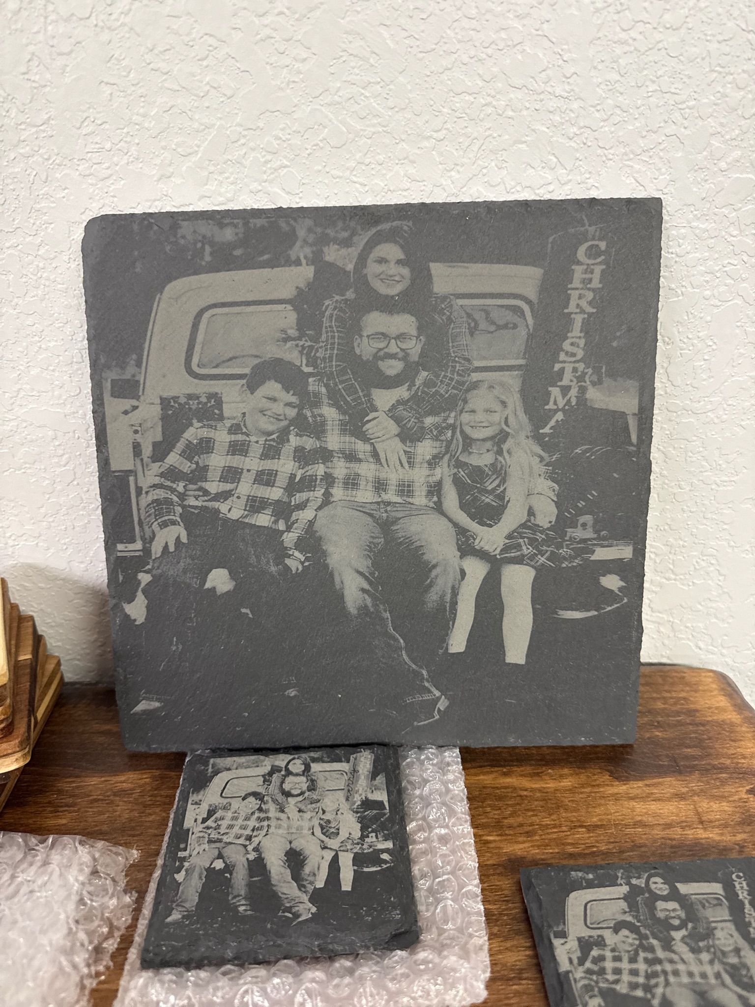 A large slate tile and two smaller coasters engraved with a family photo of four people sitting on the back of a truck.