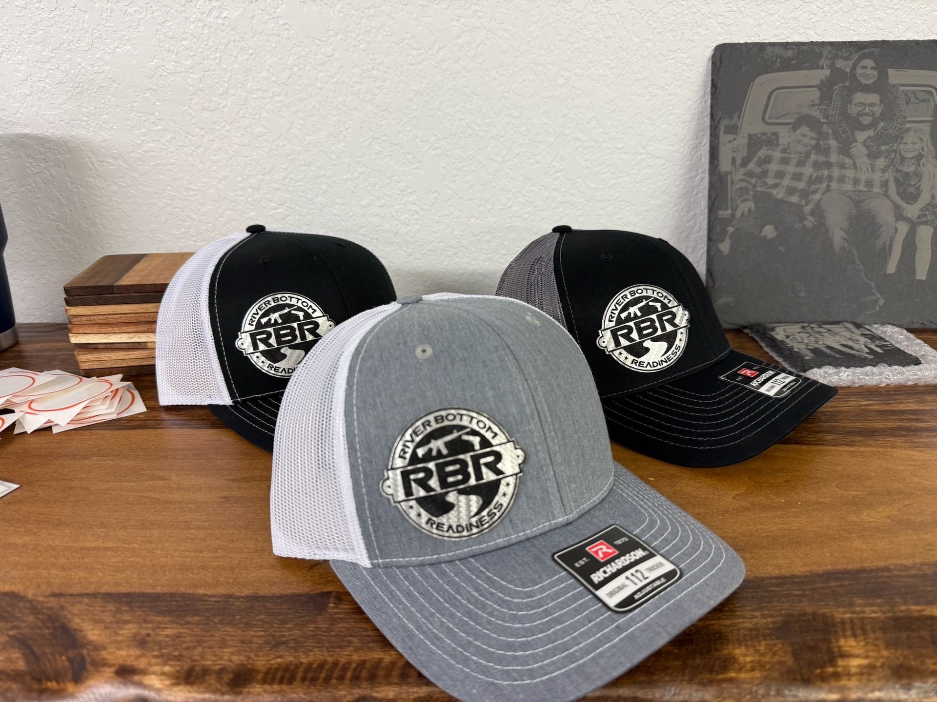 A grey and black trucker-style cap with a rectangular black leather-style patch on the front featuring stylized text.