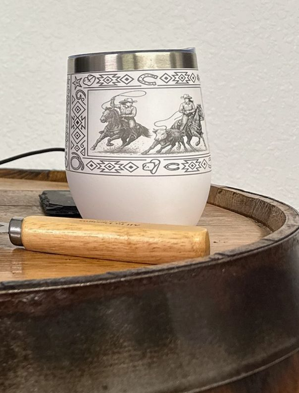 A white tumbler featuring a cowboy rope illustration sits on a wooden barrel next to a wooden-handled tool.