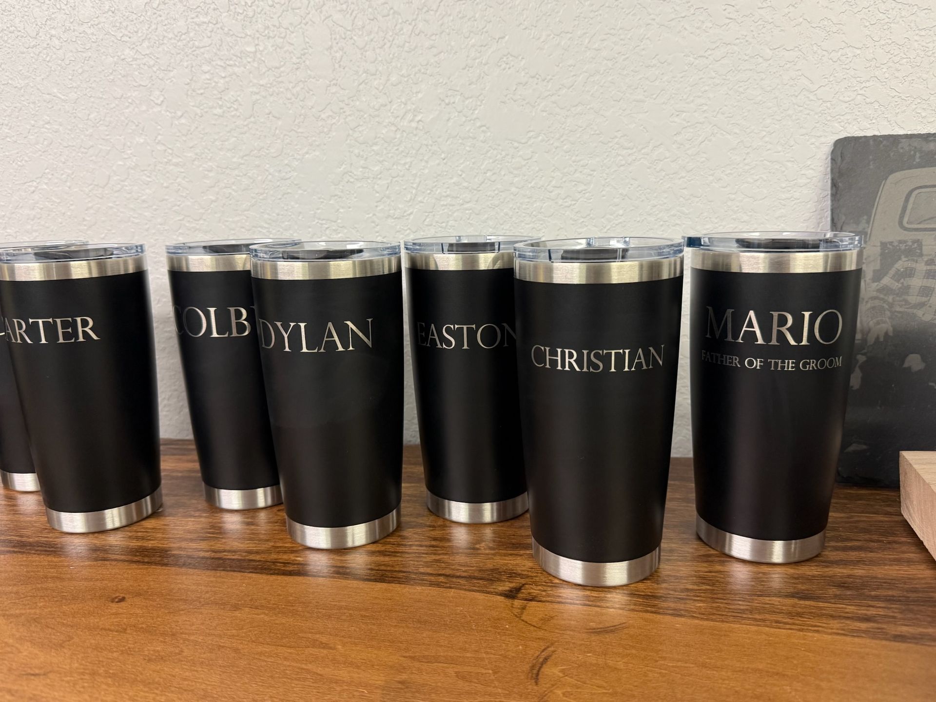 Black tumblers with silver rims engraved with names on a wooden surface.