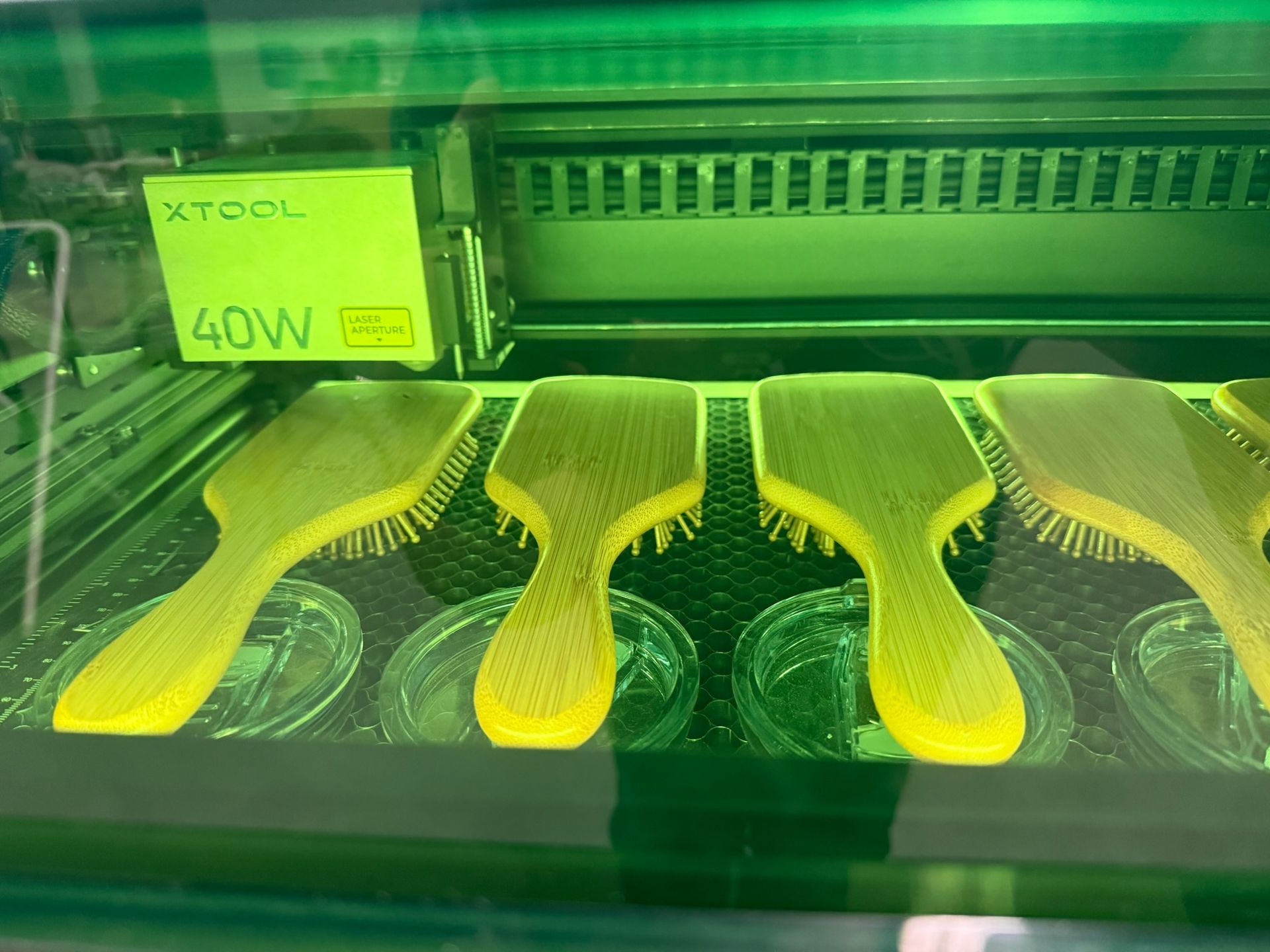 Four wooden hairbrushes arranged in a row inside an xTool laser engraving machine under bright green light.