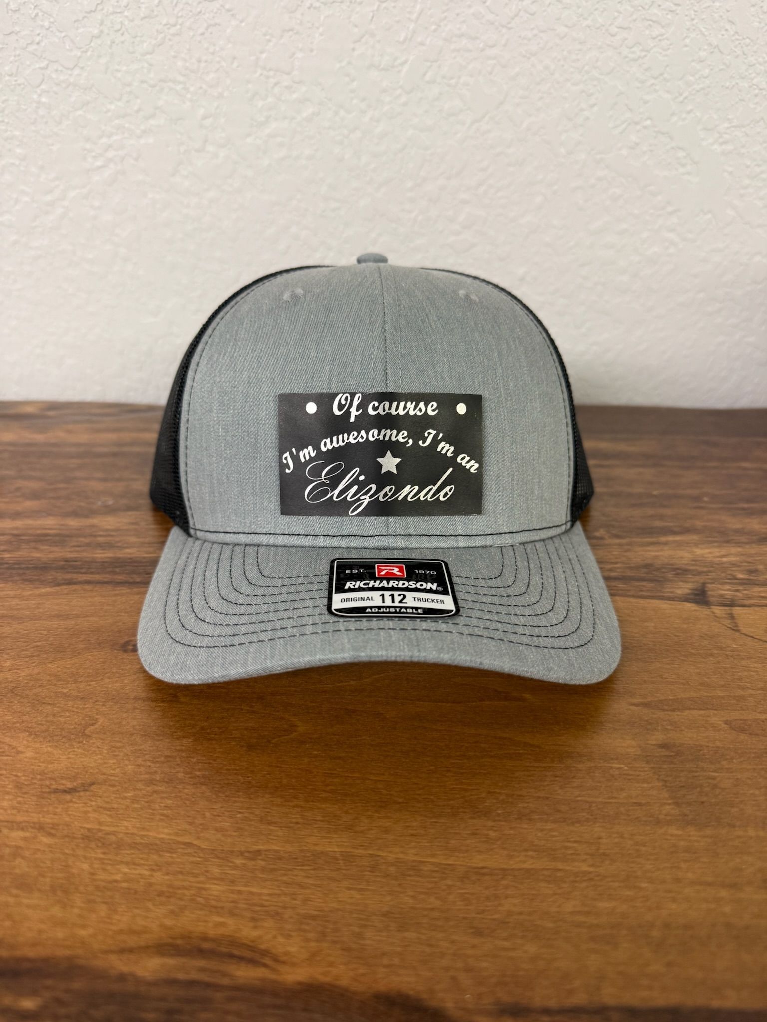 A grey and black trucker-style cap with a rectangular black leather-style patch on the front featuring stylized text.
