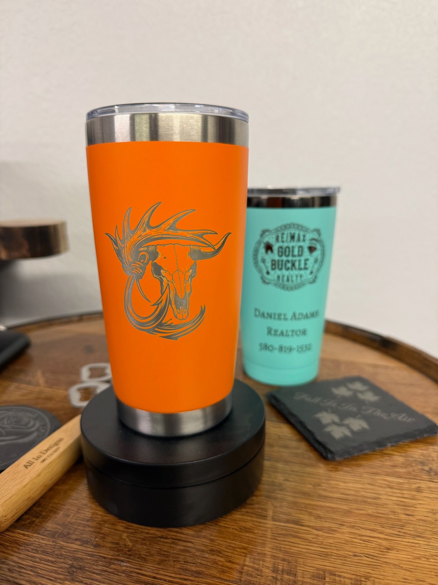 Two personalized tumblers sit on a wooden table, one orange with a bull skull design and one teal with custom text.