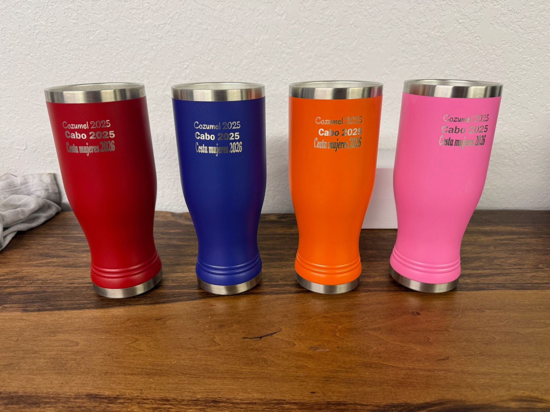 Four colorful pilsner tumblers in red, blue, orange, and pink sit in a row on a wooden surface against a white wall.