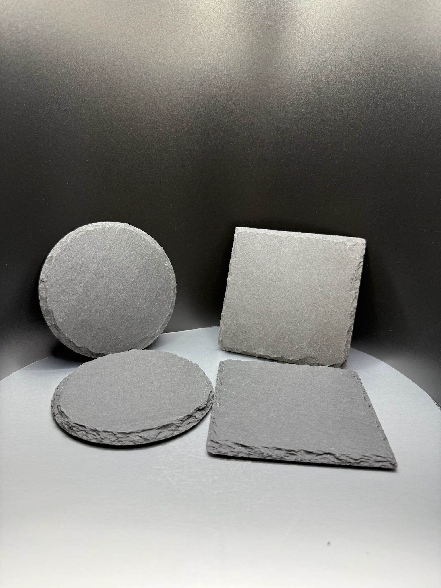 Two circular and two square dark grey stone slate coasters arranged on a reflective surface against a dark background.