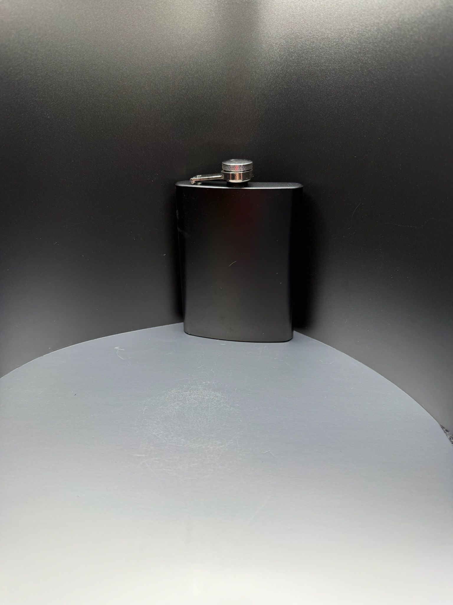 A matte black hip flask with a silver screw-top lid, centered on a light gray surface against a dark background.