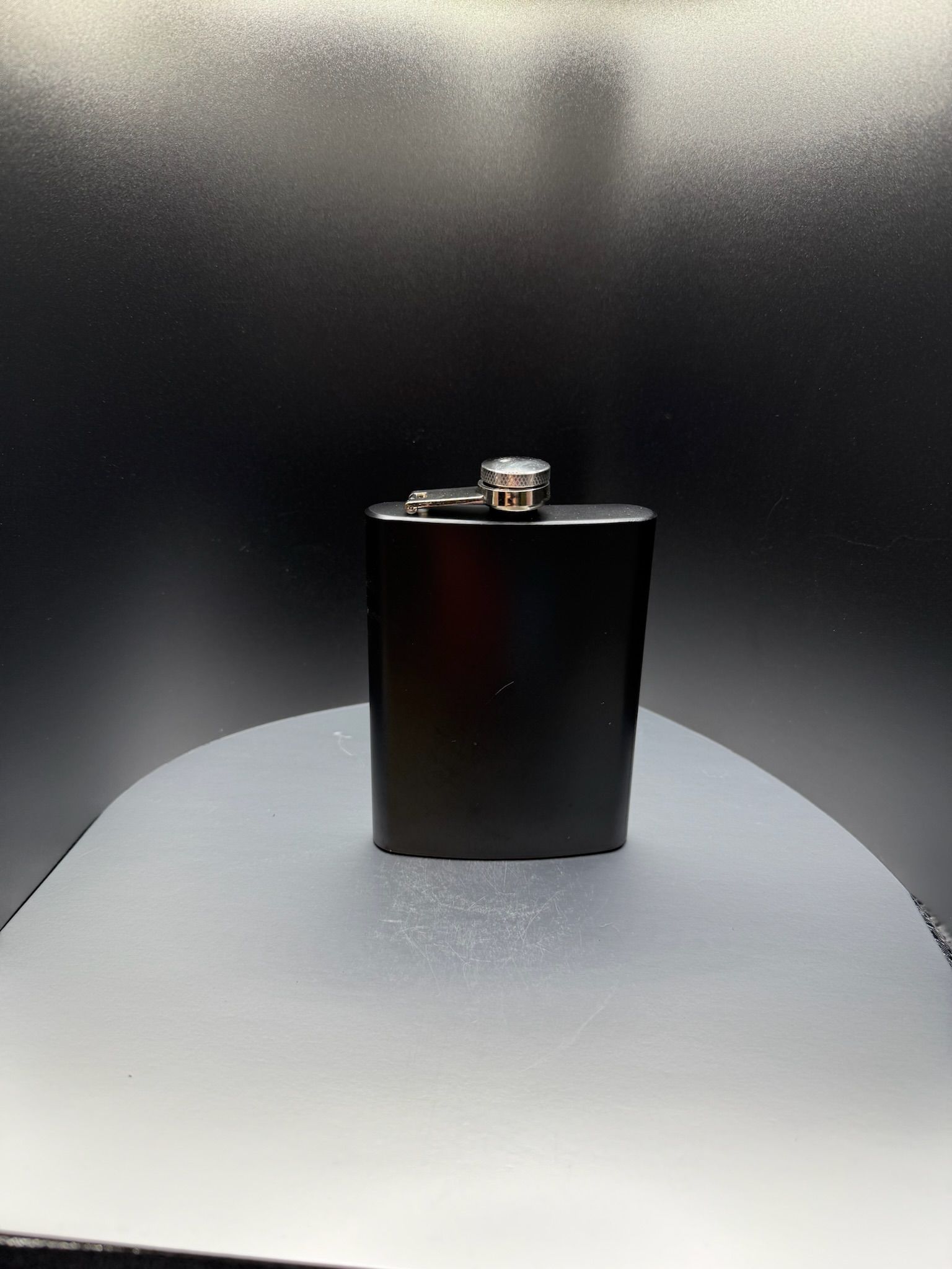 A matte black hip flask with a silver screw-top lid, centered on a light gray surface against a dark background.