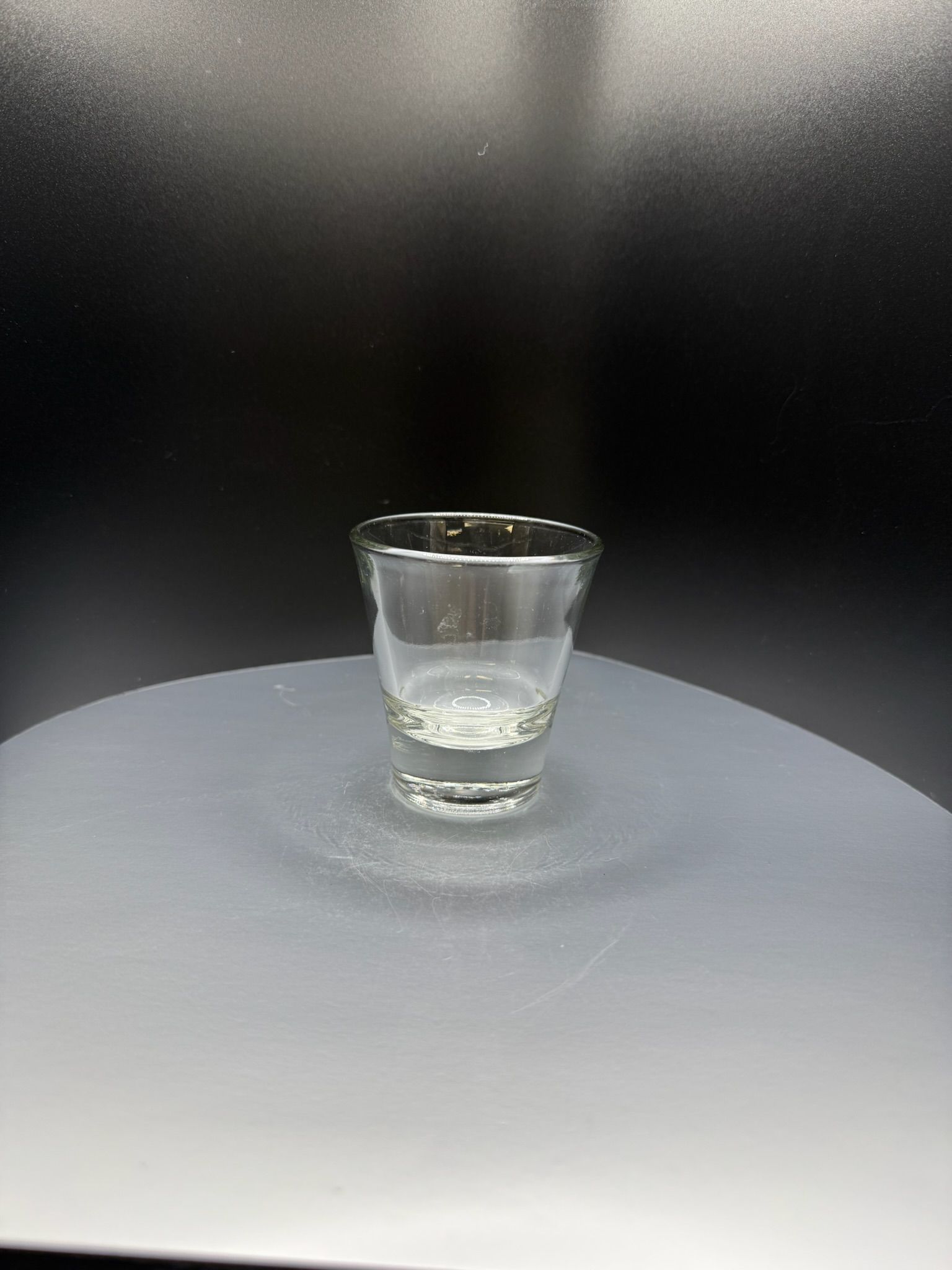 A clear glass shot glass centered on a light gray circular base against a dark background.