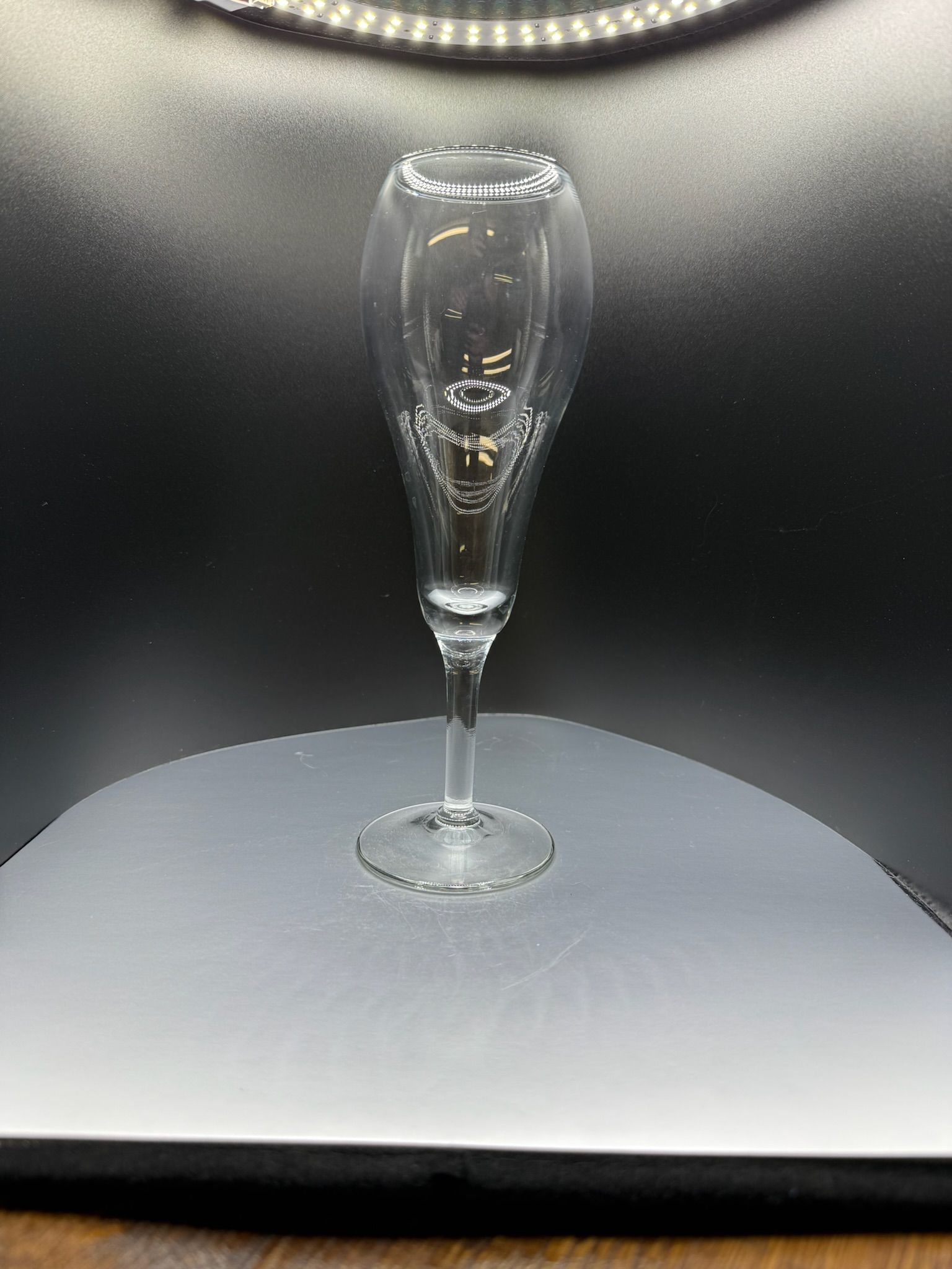 A clear, tall champagne flute stands centered on a white curved surface against a dark background.