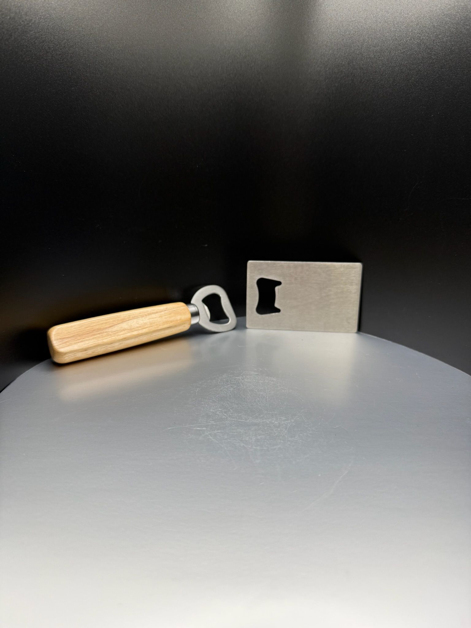 A wooden-handled bottle opener and a flat rectangular metal bottle opener sitting on a light-colored surface.
