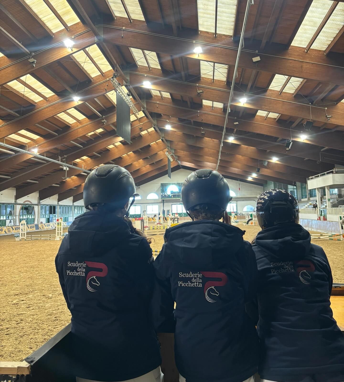 Tre persone in tenuta da equitazione assistono a un evento equestre in un'arena coperta.