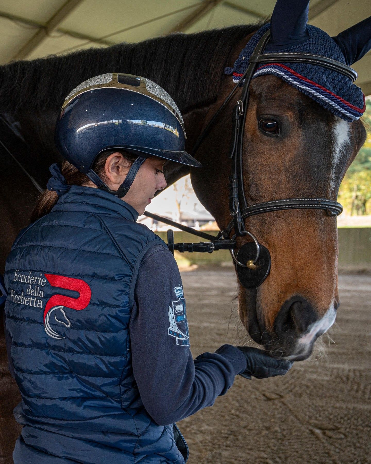 Una donna in tenuta da equitazione offre un dolcetto a un cavallo marrone, entrambi al chiuso sotto una tettoia.