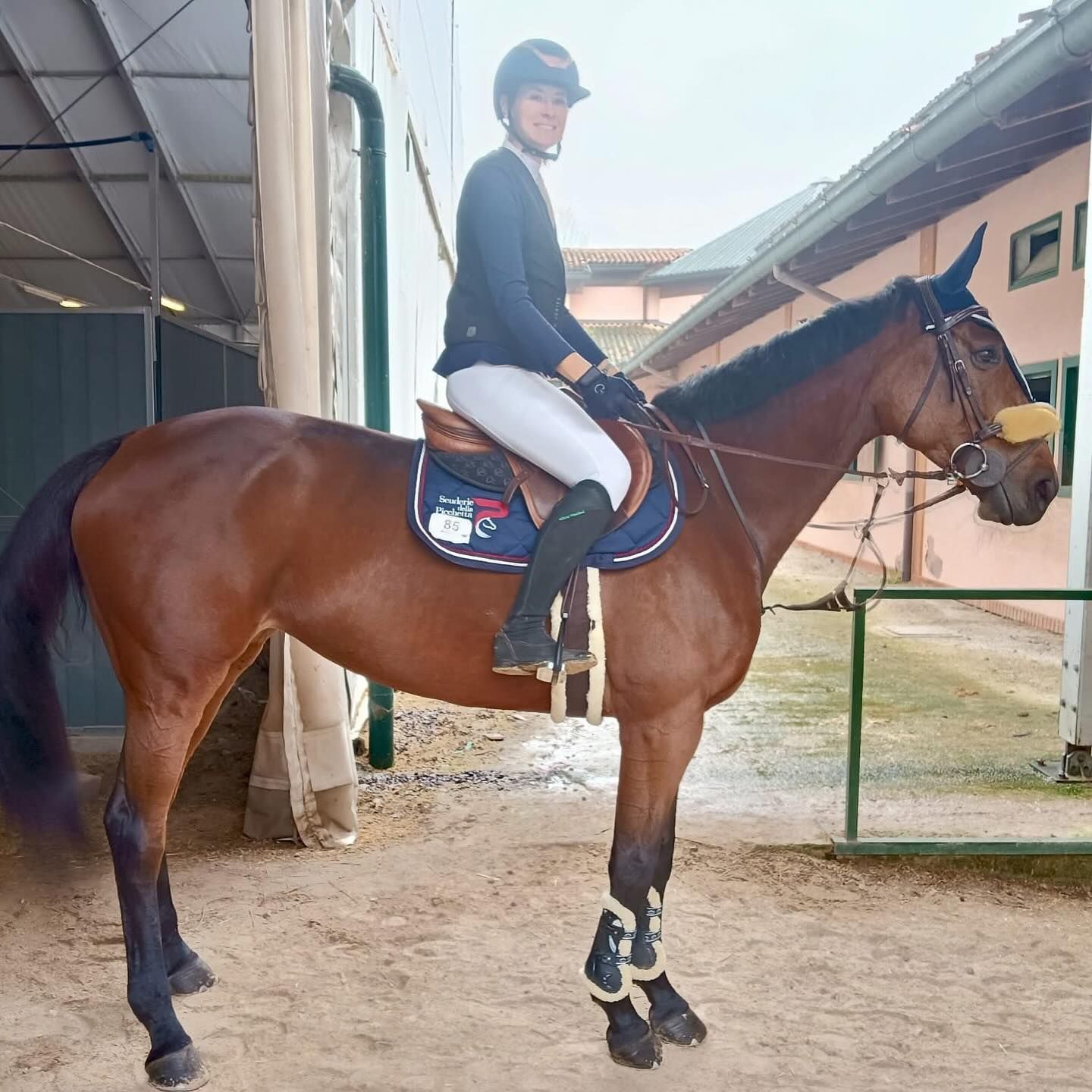 Donna a cavallo in una stalla, con casco e abbigliamento da equitazione. Il cavallo è marrone con le zampe nere.