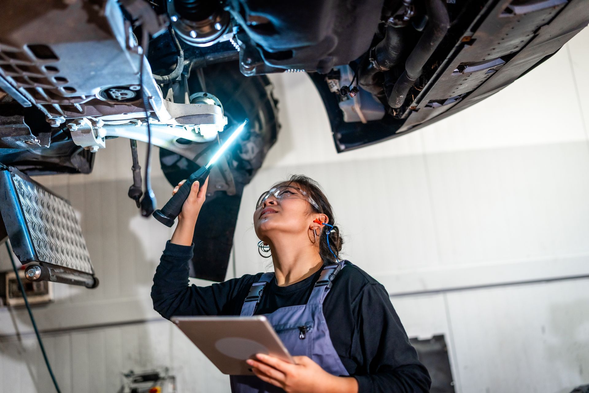Mechanic examining vehicle undercarriage with a flashlight and tablet in a garage.