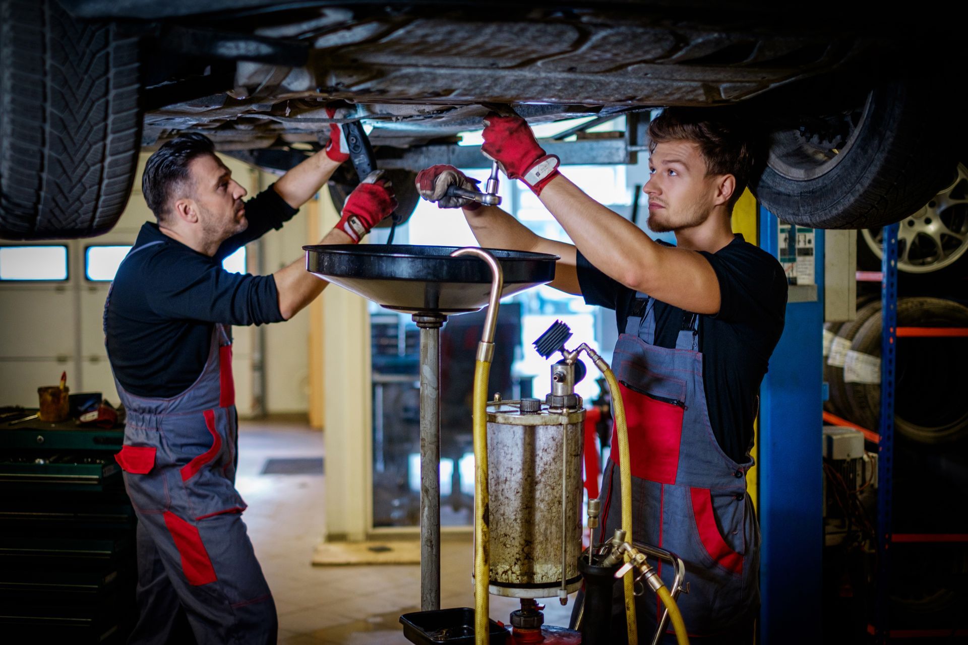 Two mechanics in coveralls working on a car from underneath, one holding a wrench, the other pouring oil.