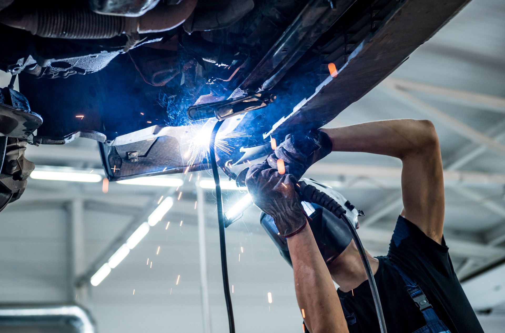 A person welds beneath a car, wearing a helmet and gloves, with sparks flying in a garage.